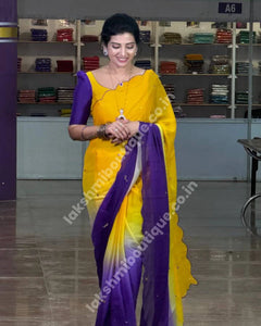 A woman modeling a yellow and purple gradient saree in a fabric showroom. The saree features a bright yellow body with a deep purple border and a matching purple pallu, with a yellow blouse.
