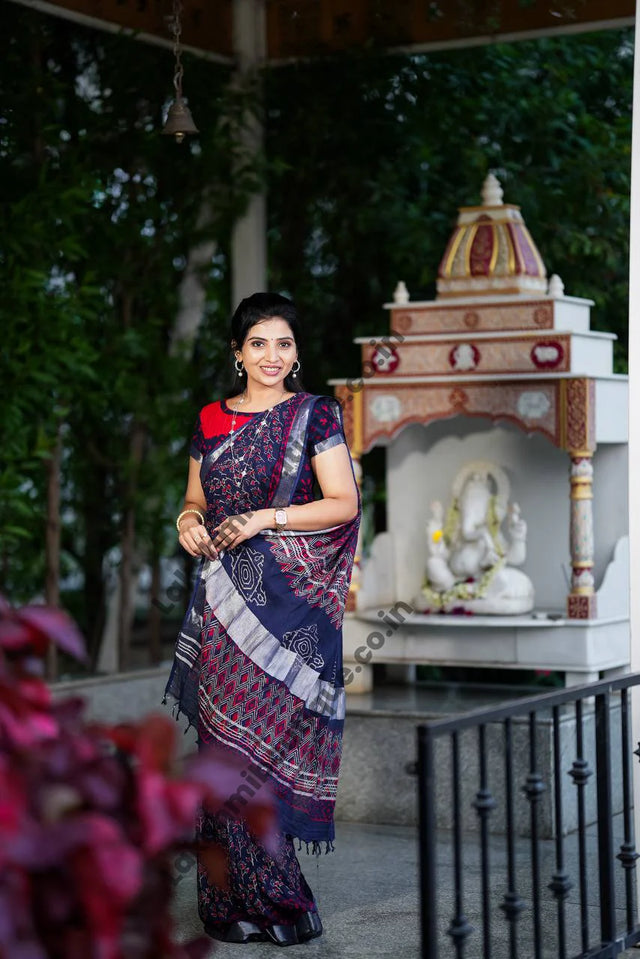 A woman wearing a navy blue and red patterned saree stands in a garden courtyard in front of a small white and red deity shrine.