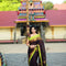 A woman wearing a navy blue and red patterned saree stands in a garden courtyard in front of a small white and red deity shrine.