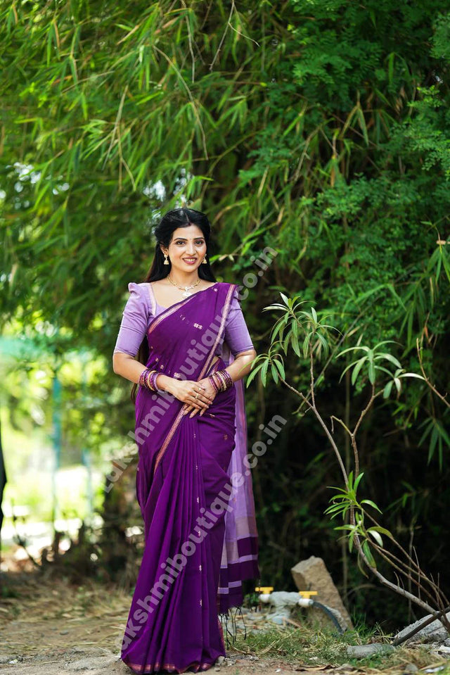 A woman wearing a navy blue and red patterned saree stands in a garden courtyard in front of a small white and red deity shrine.