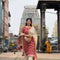 A woman wearing a red and gold patterned traditional Indian salwar kameez outfit walks toward the camera in an outdoor urban setting with a temple tower in the background.