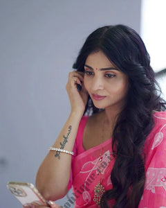 A woman wearing a navy blue and red patterned saree stands in a garden courtyard in front of a small white and red deity shrine.