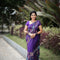A woman wearing a navy blue and red patterned saree stands in a garden courtyard in front of a small white and red deity shrine.