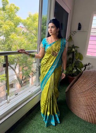 A woman wearing a bright yellow-green and turquoise blue saree stands on a balcony with a large window behind her, looking out toward green trees. The room has green artificial grass flooring, a woven chair, and indoor plants.