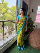 A woman wearing a bright yellow-green and turquoise blue saree stands on a balcony with a large window behind her, looking out toward green trees. The room has green artificial grass flooring, a woven chair, and indoor plants.