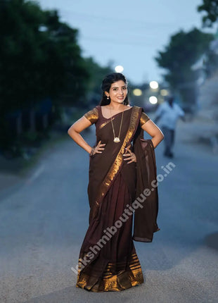 A woman wearing a dark brown and maroon saree with gold zari borders stands on a blurred road at dusk. The saree has a dark brown body, maroon pallu, and gold patterned border, with a matching dark brown blouse featuring gold embroidery.