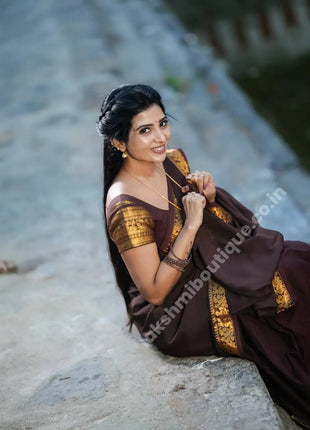 A woman wearing a dark brown saree with gold border detailing sits on a stone surface outdoors. The saree has a rich dark brown body with ornate gold patterns along the sleeve and border.