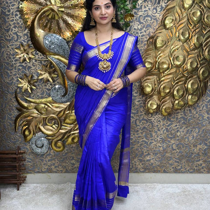 Woman in a blue saree with gold jewelry against a decorative wall.