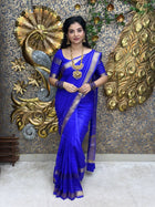 Woman in a blue saree with gold jewelry against a decorative wall.