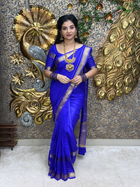 Woman in a blue saree with gold jewelry against a decorative wall.