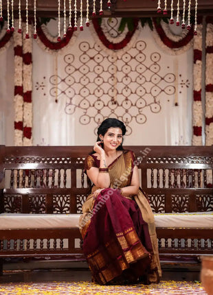 A woman wearing a traditional Indian saree sits on a carved wooden bench in a decorated indoor setting. The saree has a deep maroon body with a beige and gold border, and she is wearing a beige blouse.
