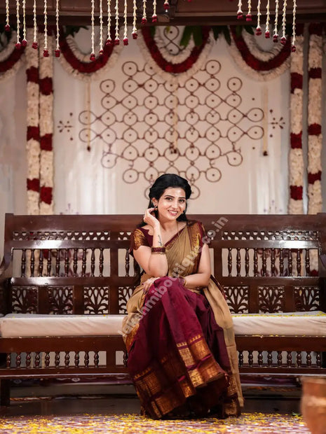 A woman wearing a traditional Indian saree sits on a carved wooden bench in a decorated indoor setting. The saree has a deep maroon body with a beige and gold border, and she is wearing a beige blouse.