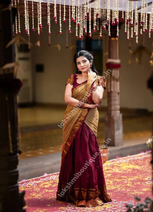 A woman wearing a traditional Indian saree stands in a decorated indoor setting. The saree has a deep maroon body with a golden-yellow border and a matching maroon blouse.