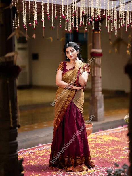 A woman wearing a traditional Indian saree stands in a decorated indoor setting. The saree has a deep maroon body with a golden-yellow border and a matching maroon blouse.