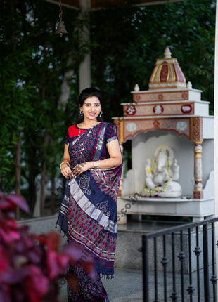 A woman wearing a navy blue and red patterned saree stands in a garden courtyard in front of a small white and red deity shrine.