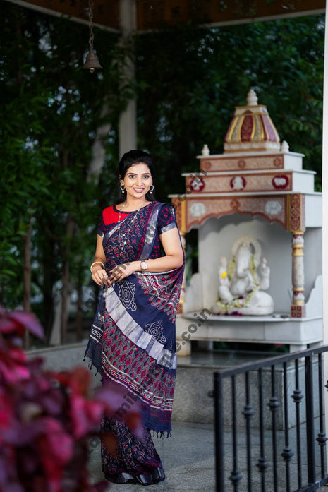 A woman wearing a navy blue and red patterned saree stands in a garden courtyard in front of a small white and red deity shrine.