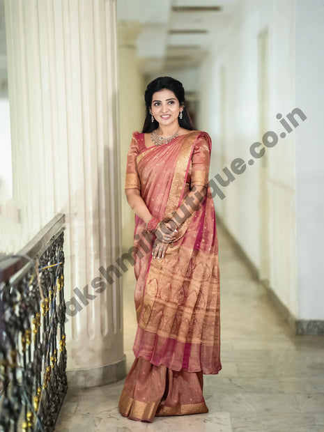 A woman wearing a traditional Indian saree stands in a hallway with columns. The saree is a dusty rose pink with gold zari patterns and borders.