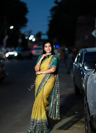 A woman wearing a yellow and green patterned saree stands on a city street at dusk, with parked cars and blurred streetlights in the background.