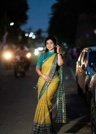 A woman wearing a yellow and green patterned saree stands on a street at night with cars and motorcycle lights blurred in the background.